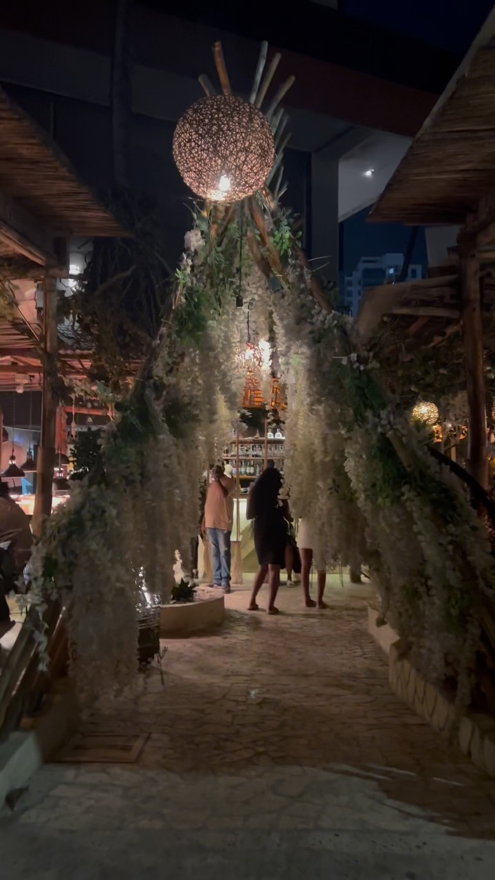 Entrance to a rooftop bar at night under hanging white flowers and woven lamps